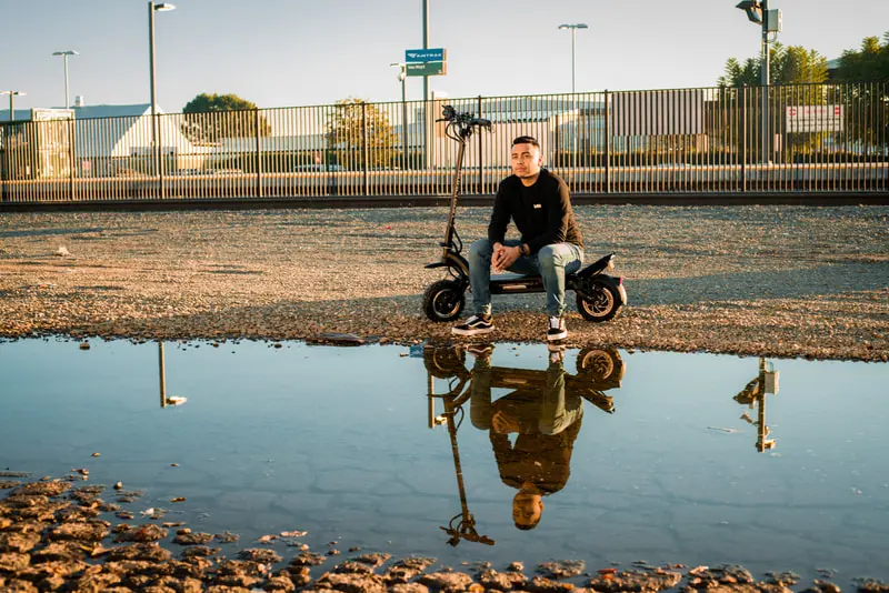 Rider and electric scooter reflected in a shallow puddle.